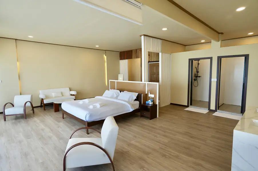 A spacious suite at F Lina Water Villa Resort near Semporna featuring a large bed, modern white armchairs, and rustic timber floor with smooth wooden boards, cooled by air conditioning.