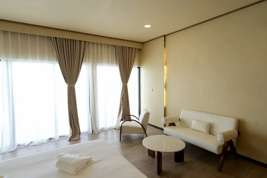 A bright bedroom at F-Lina Water Villa Resort near Semporna showcasing a modern seating area facing panoramic glass doors, smooth wooden boards, and air conditioning.