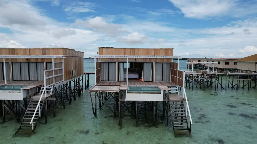 A modern local style overwater chalet painted white featuring a sloped roof and a spacious wooden deck with a shaded timber pergola at F-Lina Water Villa Resort.