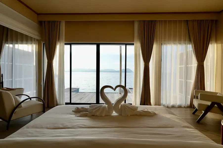 A spacious bedroom featuring a comfortable bed with towel swans and stylish armchairs, accented by sliding glass doors that open to a balcony with a sweeping seaview over the Celebes Sea, embodying elegant coastal design at F-Lina Water Villa Resort.