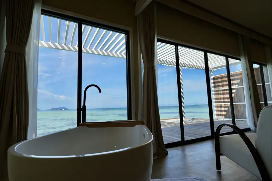 A modern white soaking tub with a matte black faucet and wooden tray overlooking the clear sea through expansive glass doors at F-Lina Water Villa Resort.
